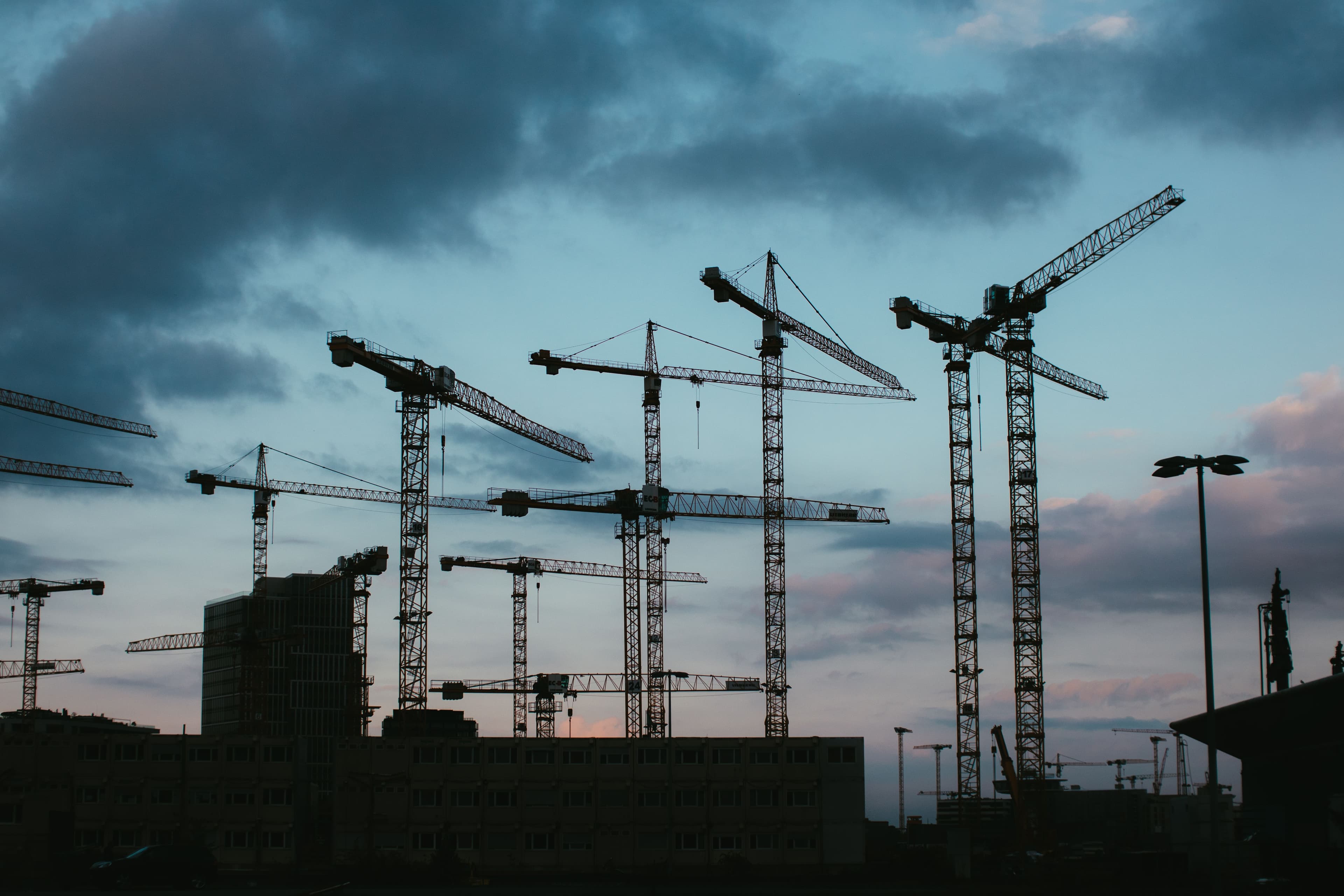 Multiple tower cranes silhouetted against a dusk sky at a large construction site