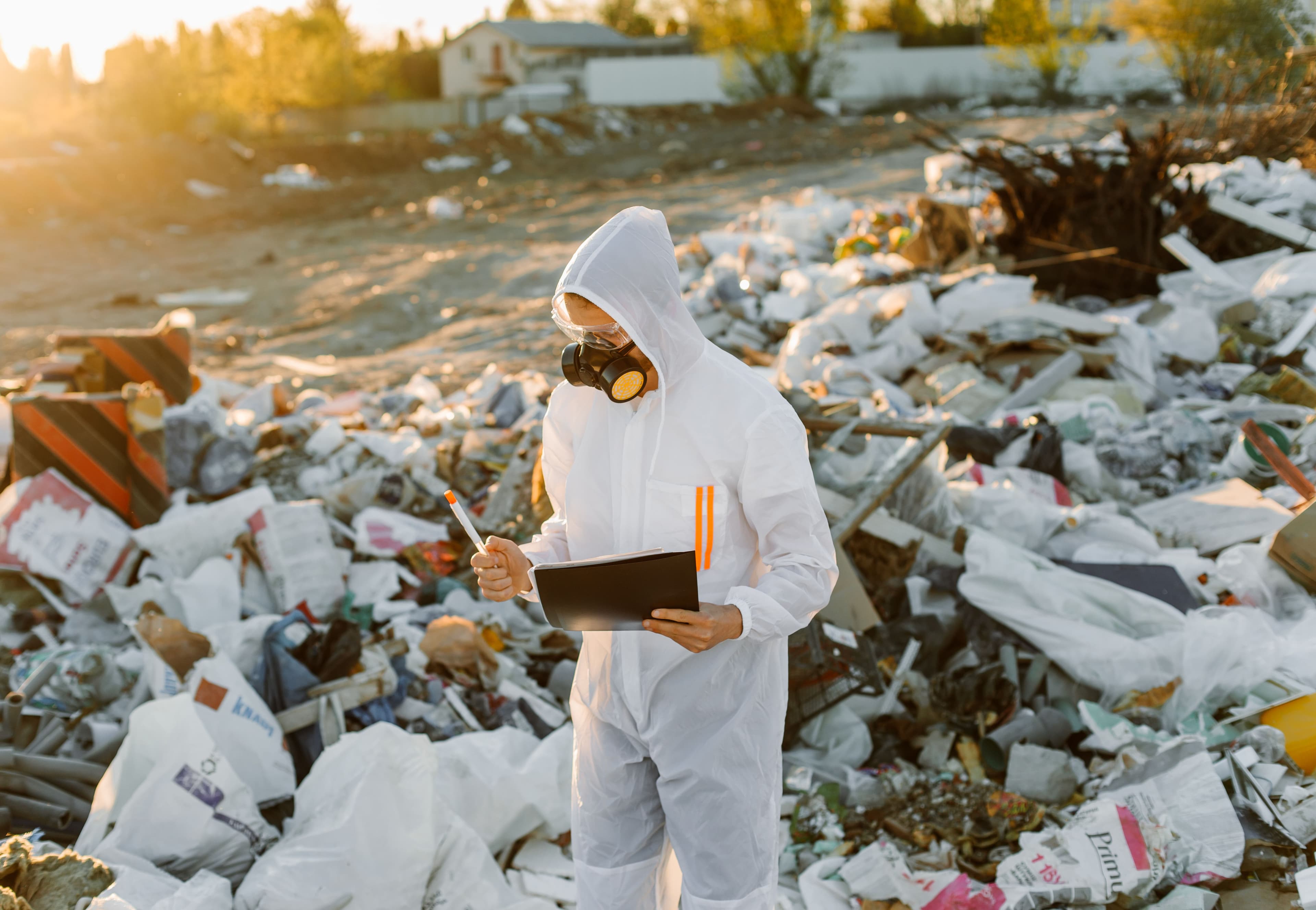 Worker in a protective coverall and respirator inspecting a waste site with a clipboard