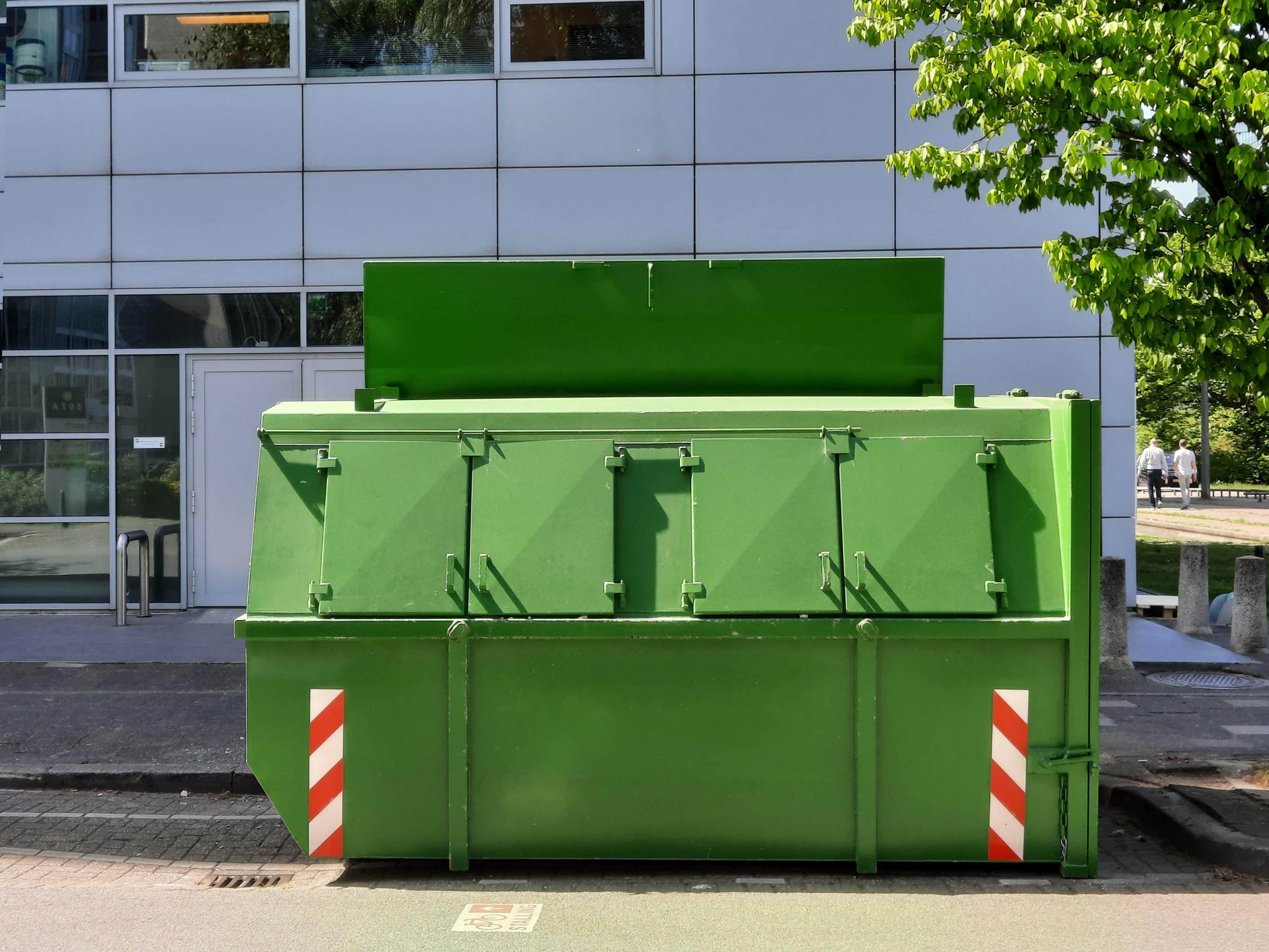 Large green waste compactor container on a street outside an office building