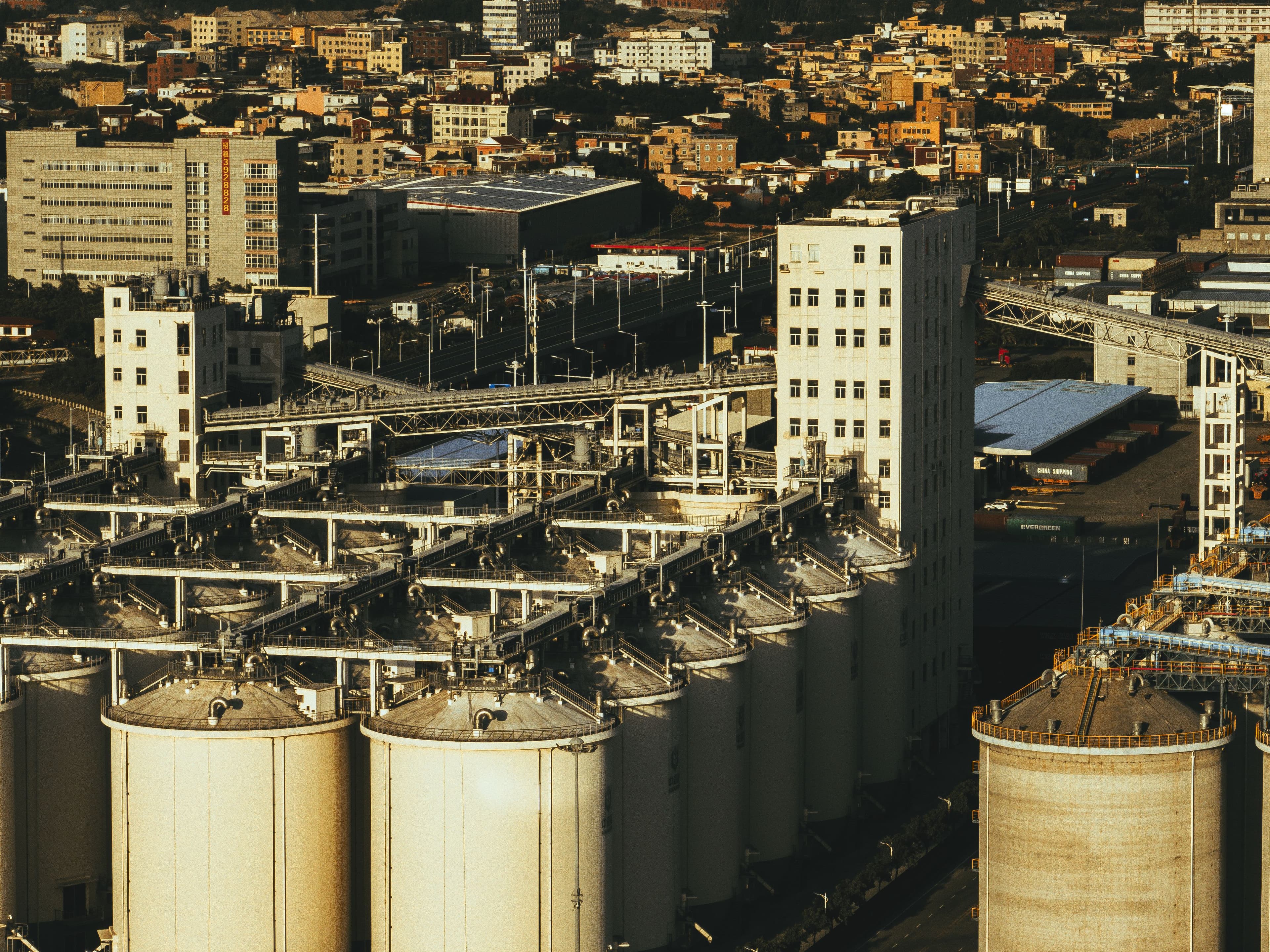 Aerial view of industrial storage silos, conveyor gantries and shipping containers in a dense port-city district